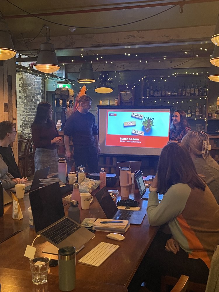 Members of RKH sit around a table looking up at a screen, members of 72Point Group stand next to the screen delivering training